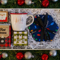 Gift set with mug, bath bomb, and hot cocoa mix on a bed of grass with Christmas ornaments.