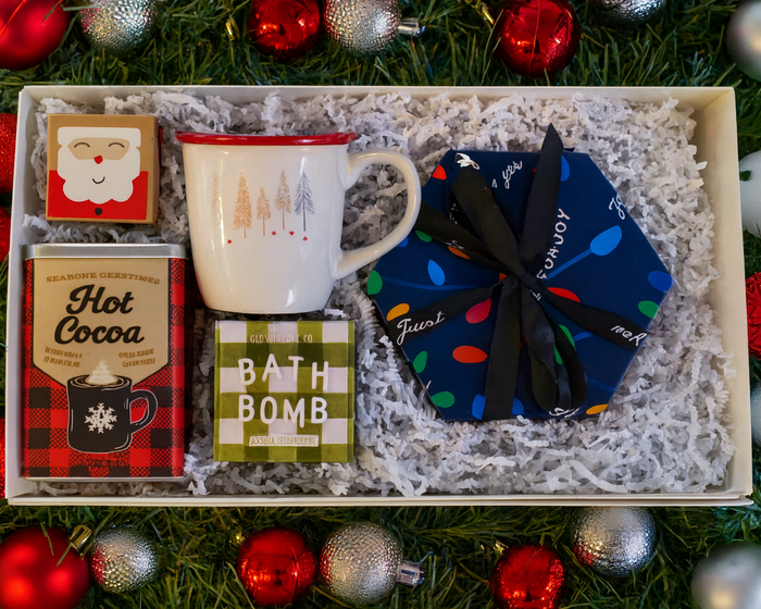 Gift set with mug, bath bomb, and hot cocoa mix on a bed of grass with Christmas ornaments.